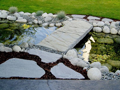Water feature in a residential yard with large slate stepping stones, a bridge, and river rock lining a small creek